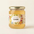 A glass jar of Linden honey with a gold metal lid and a colorful floral label, set against a plain off-white background.